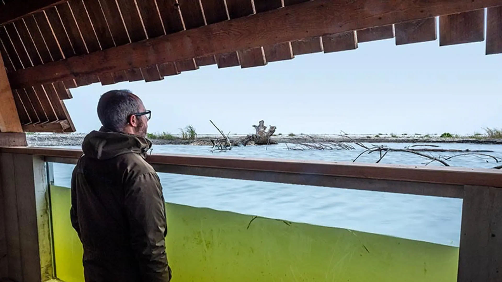 Man in a wooden building structure looking out into a half-immersed lake.