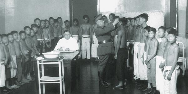 Black and white photograph of children aligned around a doctor.