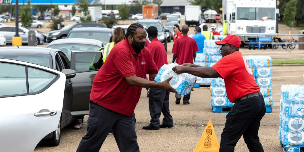 A man giving a pack of water to another man. Both are wearing red.