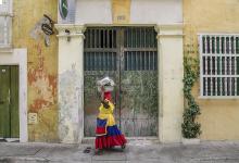 Women carrying a basket on head, walking in front of green gate in a Columbian neighborhood.