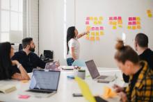 Woman pointing to sticky notes and others sitting at a table with laptops out