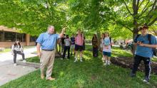 Students taking their class under green trees, the professor John Sprinkle is pointing.