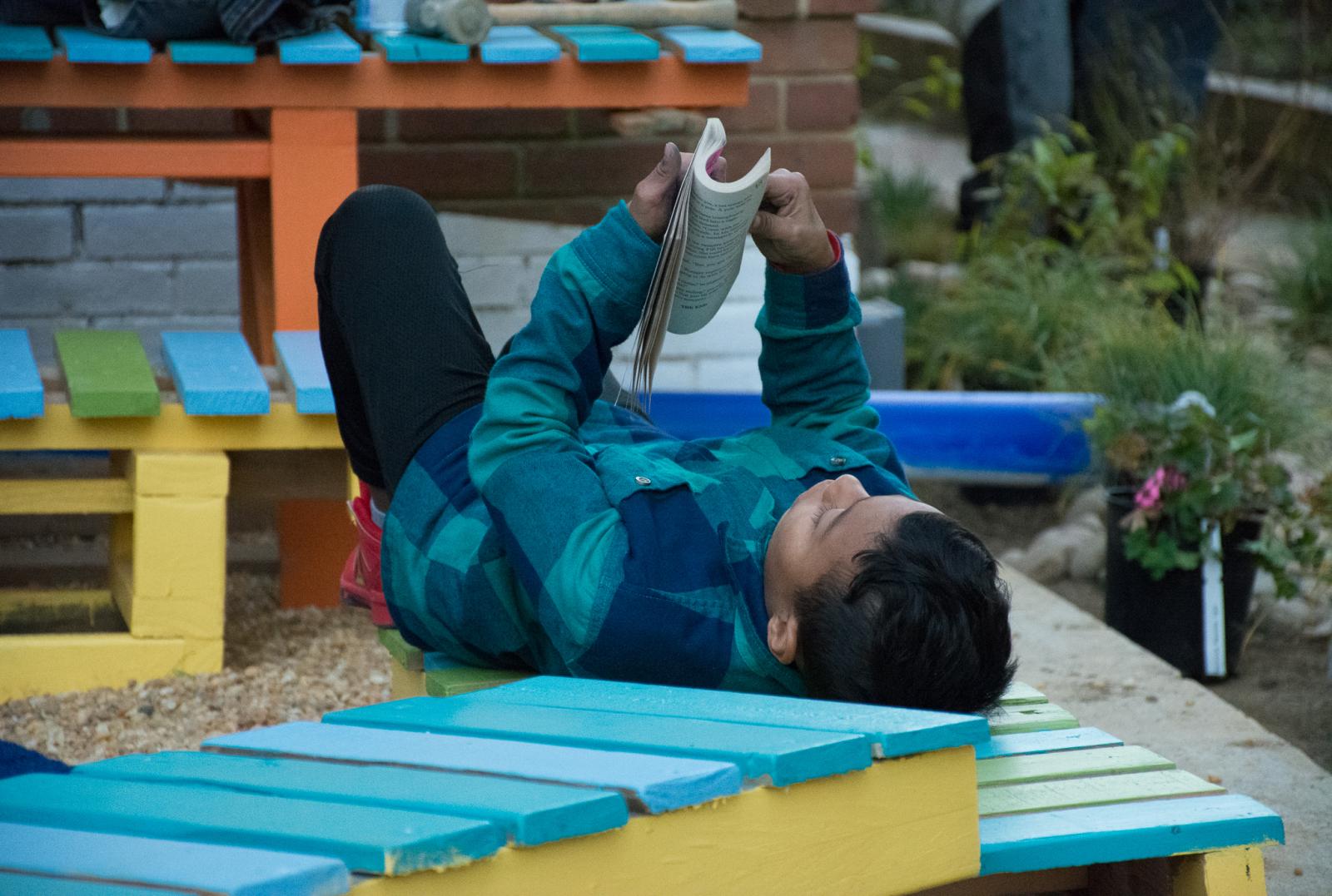 Boy reading while laying on a colorful blue and yellow bench at the Lightscape Rain garden