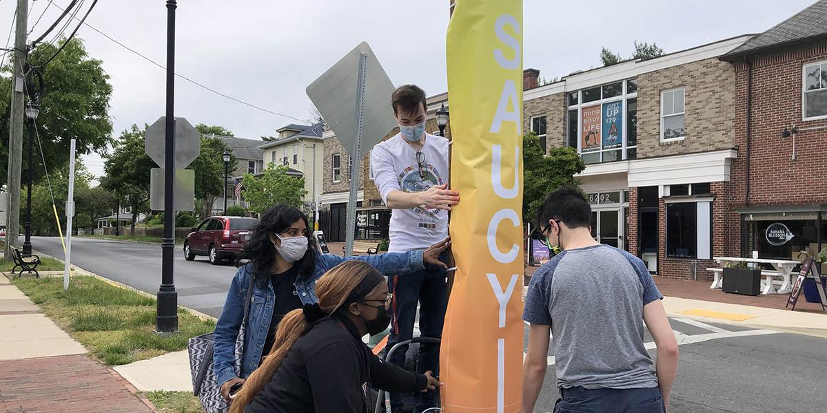 Students hanging a lamp post sign that reads "Saucy" for the "Taste of Riverdale" project.