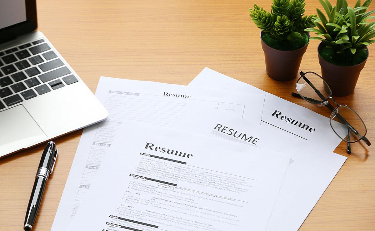 Resumes on a desk with a fake plant and laptop.